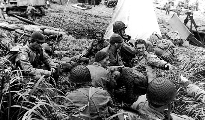 Men of the First Regiment command group huddle on Beach 9-Blue on D-Day 15 August 1943. The white triangular shape behind the men is a beach marking panel erected to guide the subsequent waves of troops to the beach.